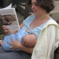 A pale skinned woman reads 'Breastfeeding: A Parent's Guide' while nursing a baby