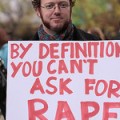 A man with dark ginger hair and whiskers is holding a sign which says BY DEFINITION YOU CAN'T ASK FOR RAPE