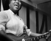 Black woman in white blouse singing, with electric guitar.