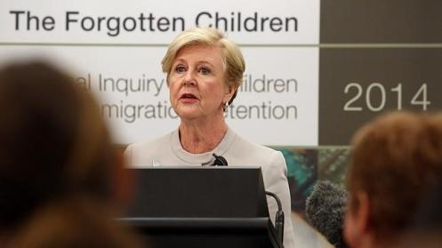 Short-haired white woman at lectern in front of sign reading 'Forgotten Children'.