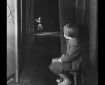 B&W photo of a toddler girl on a stool watching a woman on stage in the distance.